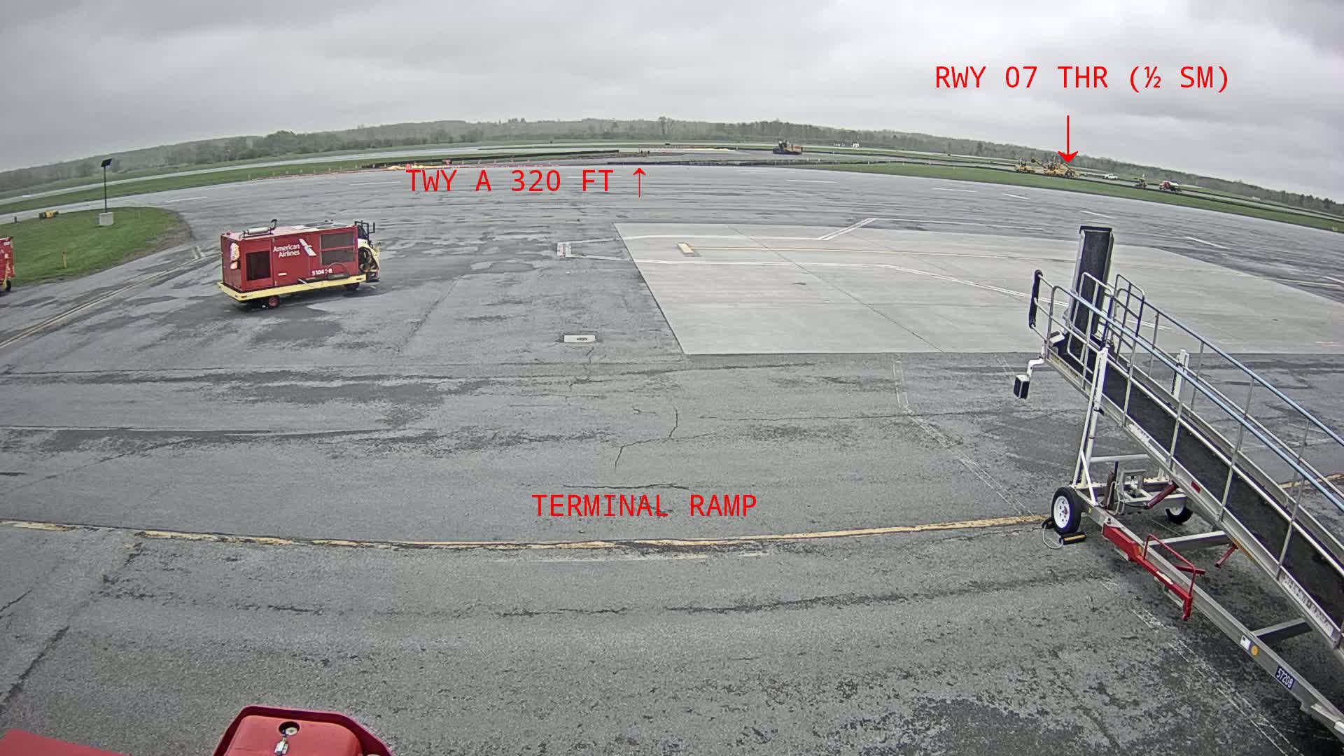 A tarmac view on an overcast day shows a red baggage tug, a jetway, and some distant construction equipment.