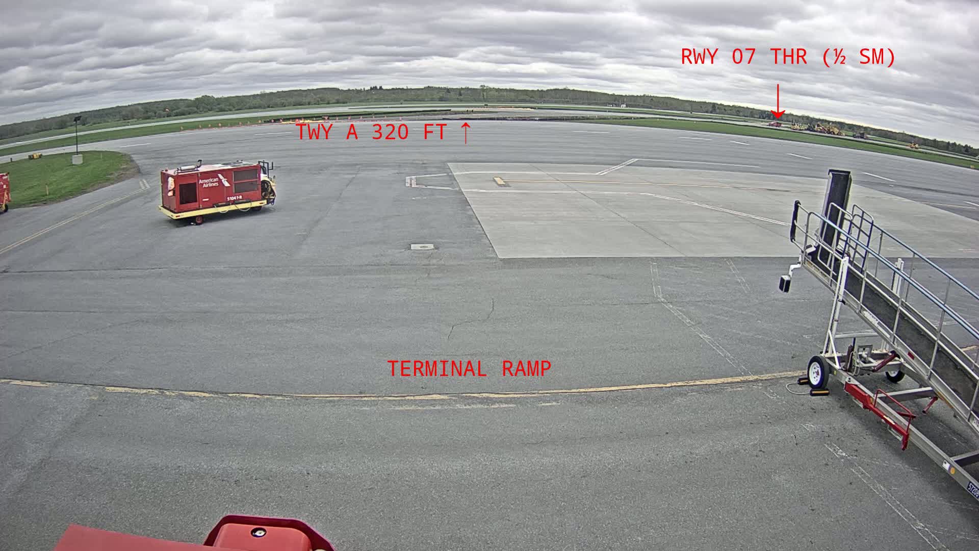 An overcast day at an airport shows a tarmac with a baggage tug, jet bridge, and a view of a runway in the distance.