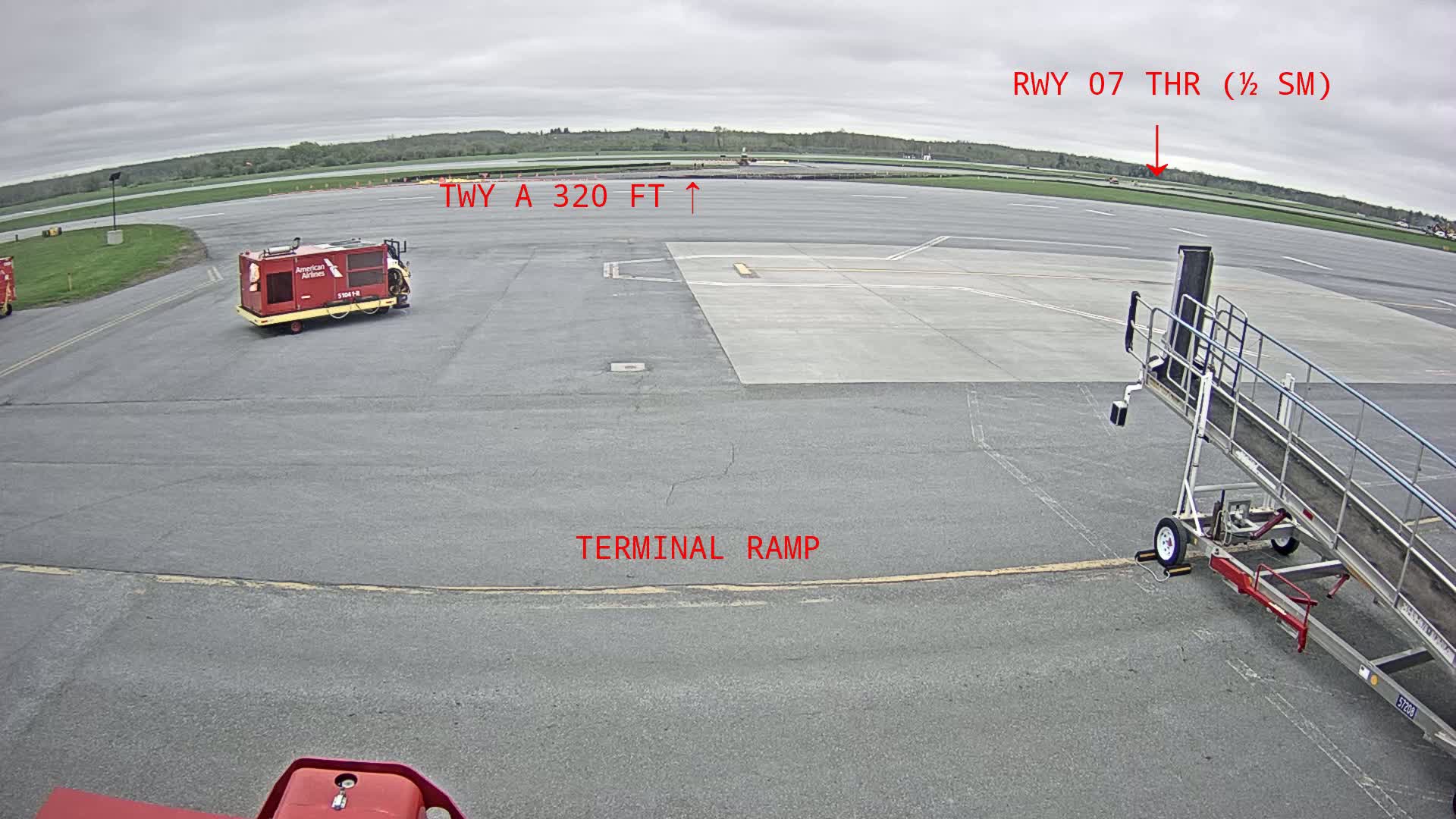 A terminal ramp at an airport on an overcast day shows a baggage tug and a jet bridge, with a runway visible in the background.