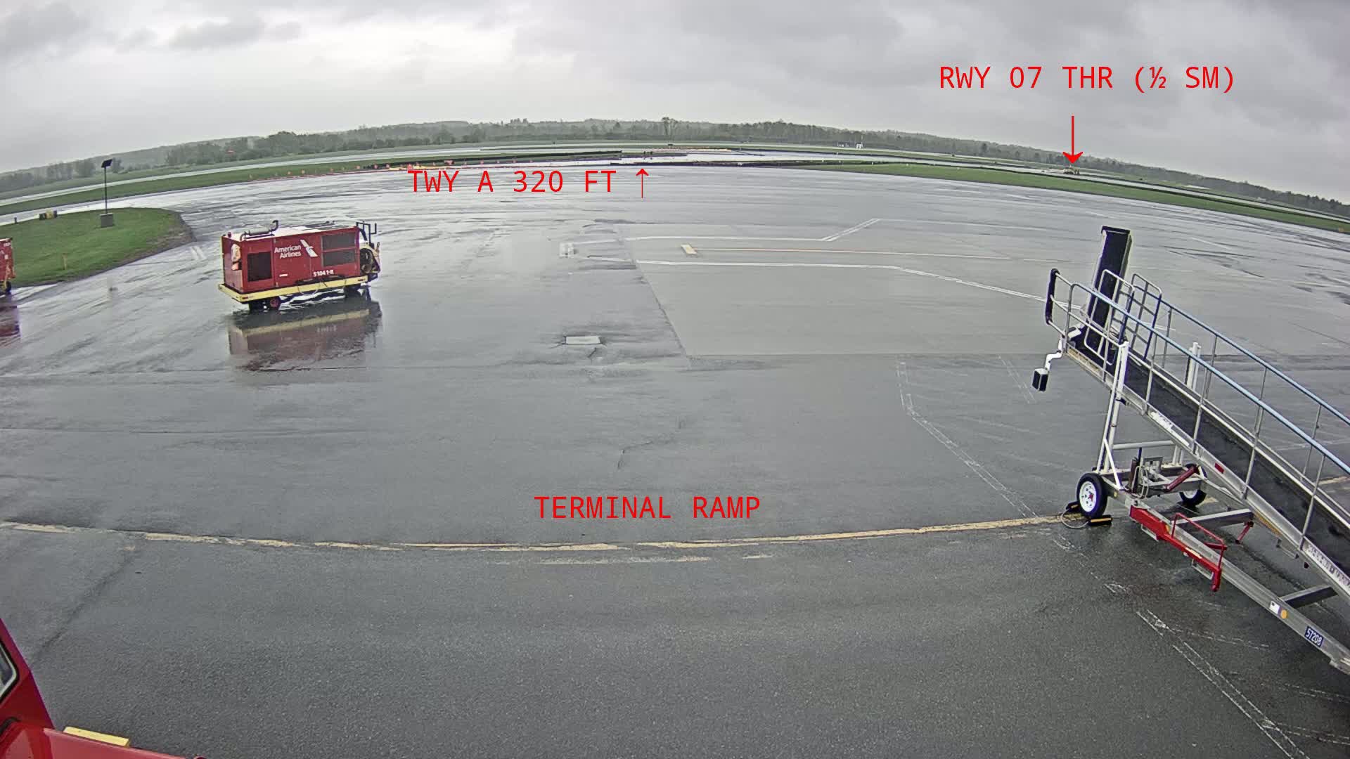 A wet airport tarmac is seen, with a jet bridge, a service vehicle, and a portion of a runway in the distance under an overcast sky.