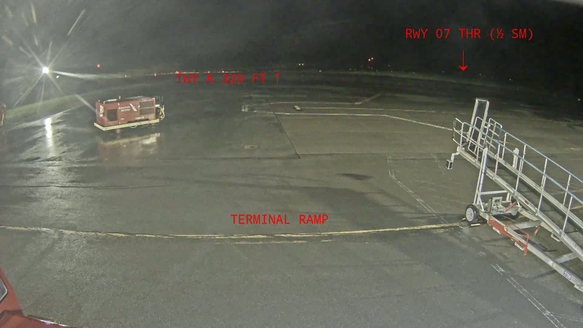 A wet airport tarmac at night shows a baggage cart, a jet bridge, and runway lights.