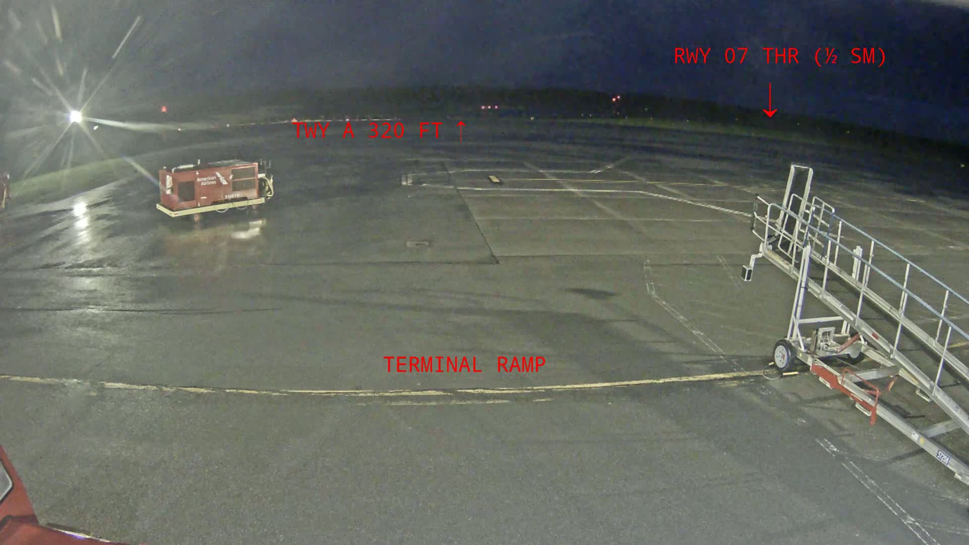 A wet airport tarmac at night shows a baggage cart, a jet bridge, and runway lights in the distance.