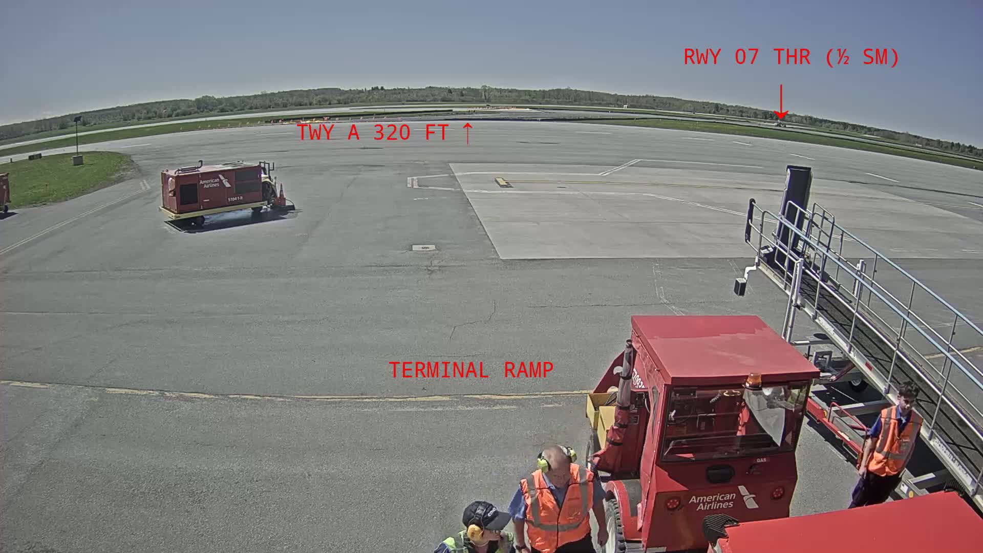 A tarmac on a sunny day shows several ground service vehicles near a jet bridge, with a runway and grassy area visible in the distance.