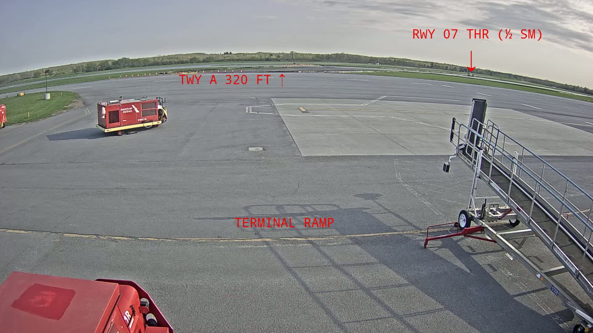 A tarmac view shows a red baggage cart, a jet bridge, and a runway under a mostly clear sky.