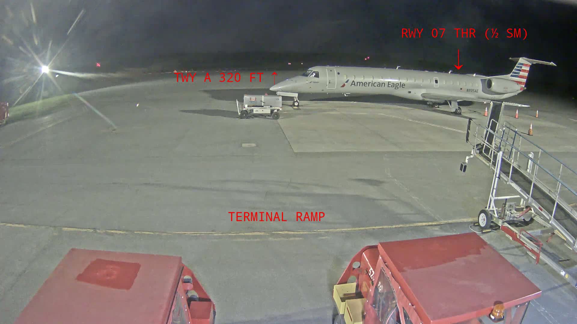 At night, an American Eagle airplane is parked on an airport tarmac near a jet bridge and ground support vehicles.