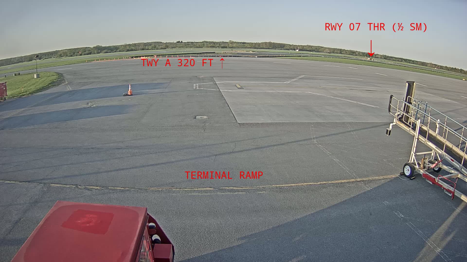 A tarmac view shows an airport terminal ramp, a jet bridge, and a portion of a runway visible in the distance under a clear, bright sky.