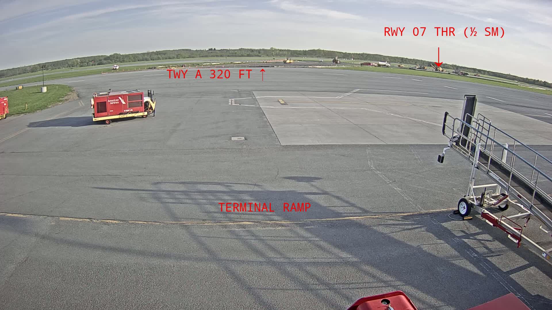 A mostly sunny tarmac shows airport ground service equipment near a terminal ramp with a runway in the distance.