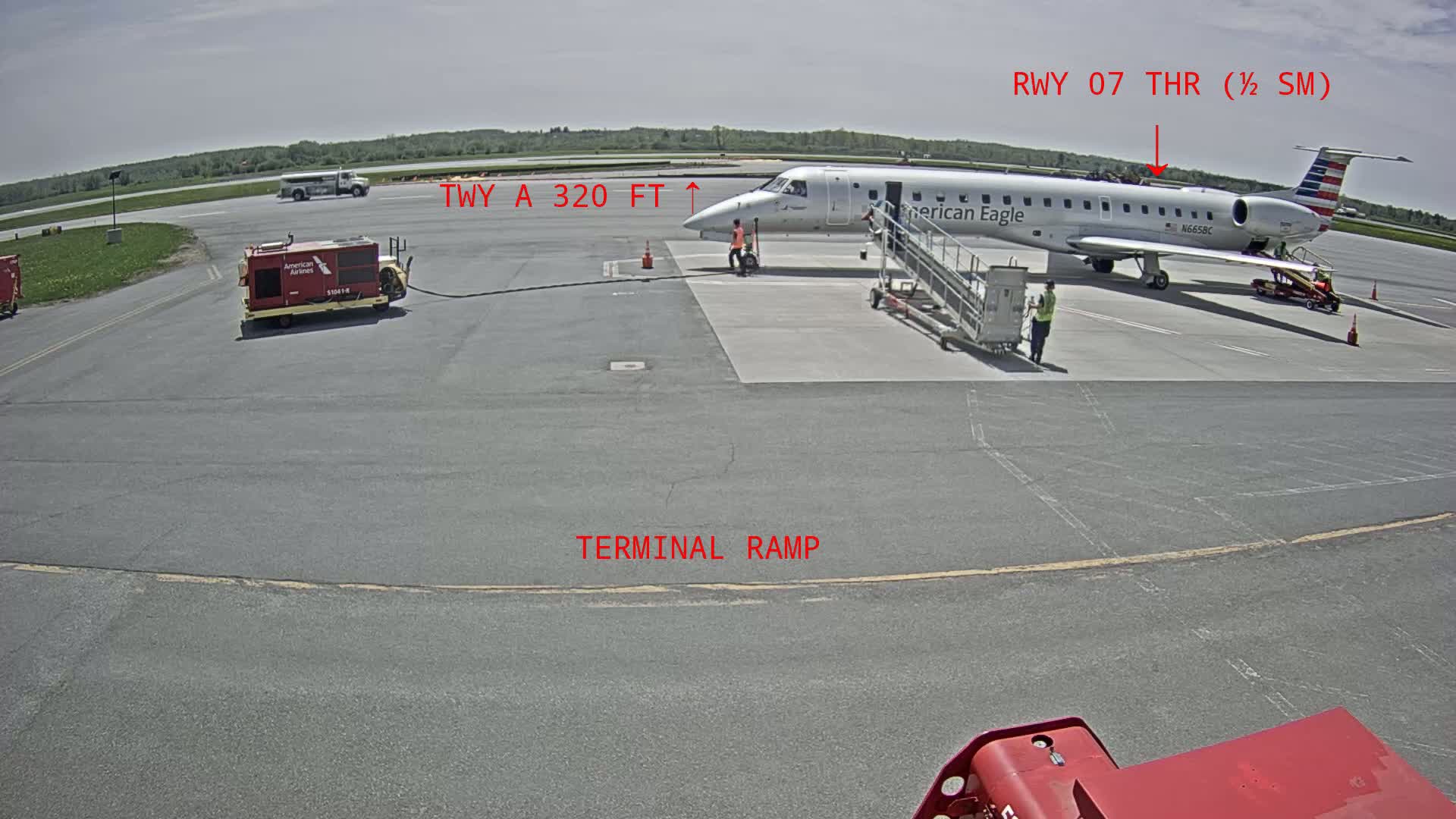 On a sunny day, an American Eagle regional jet is parked at a terminal ramp on an airport tarmac, with ground support equipment nearby.