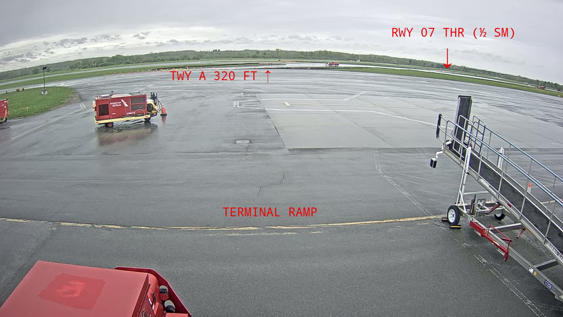 A wet airport tarmac is viewed from a low angle, showing a terminal ramp, airport service vehicles, and a jet bridge.