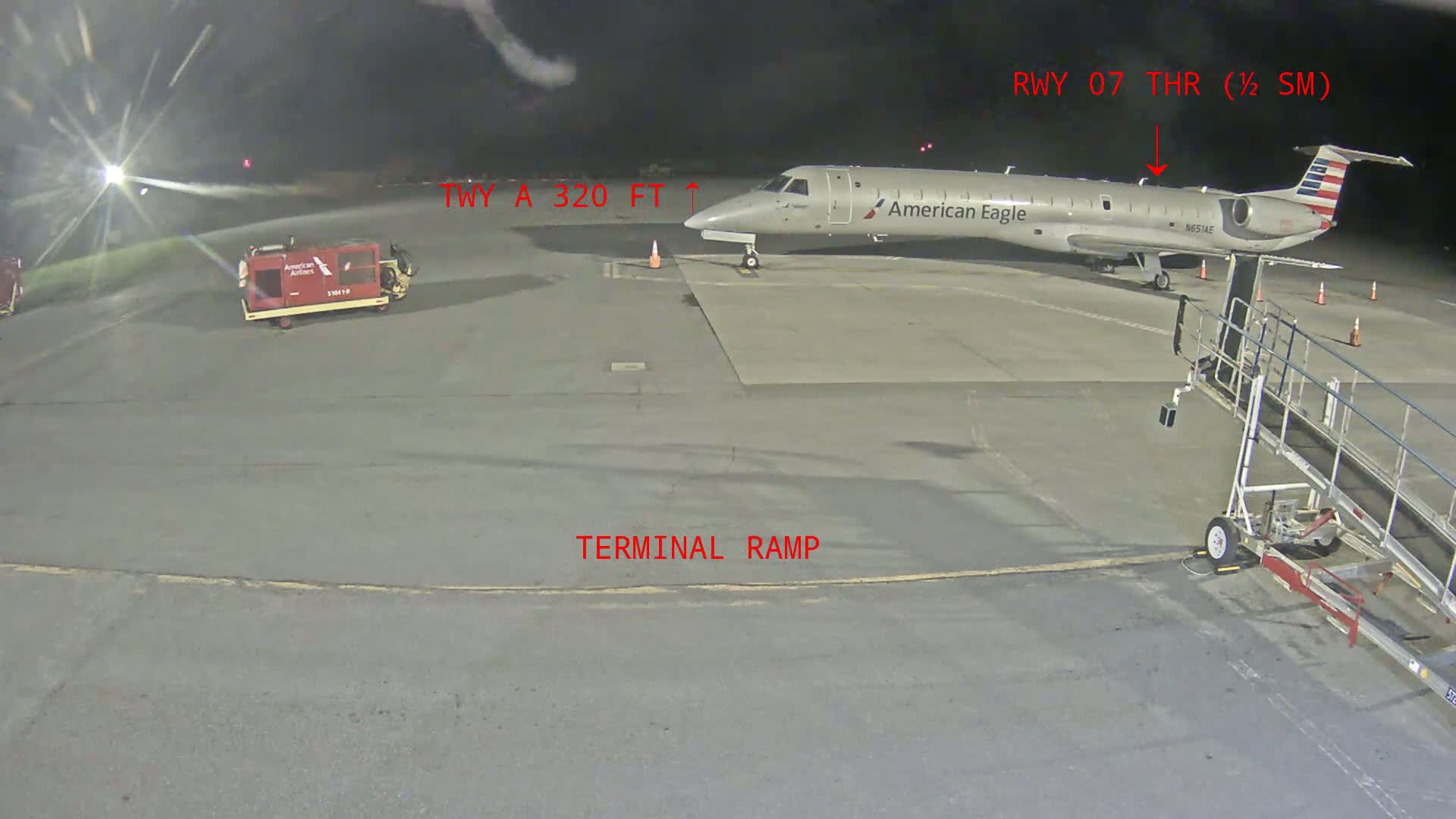 At night, a silver American Eagle regional jet is parked at a terminal ramp near a baggage tug and jet bridge.