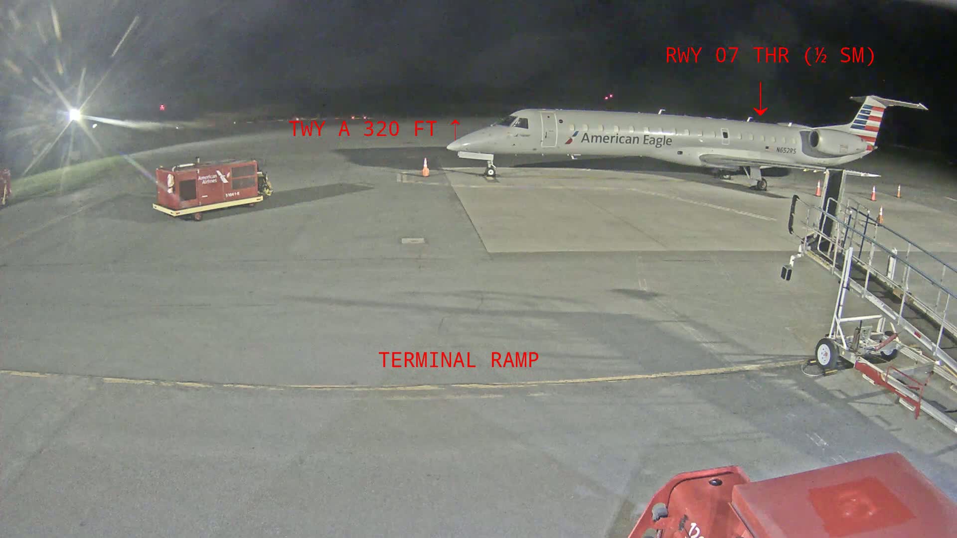 At night, an American Eagle jet is parked on a tarmac next to a jet bridge, with airport ground support equipment nearby.