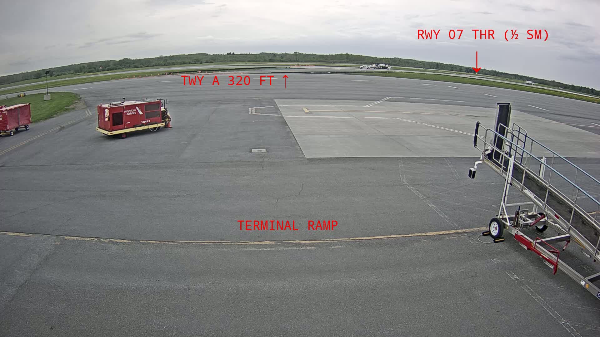 An airport tarmac under overcast skies shows a baggage cart, a jet bridge, and other airport ground support equipment.