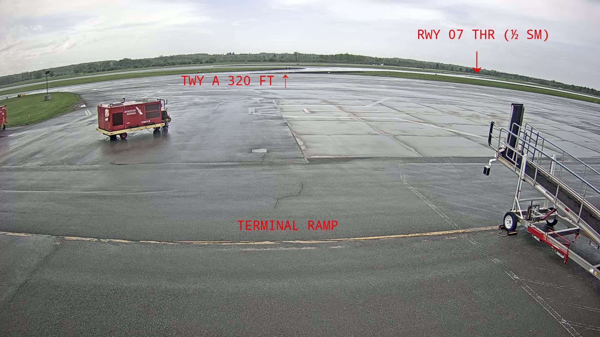 A wet airport tarmac shows a baggage cart, a jet bridge, and distant runway.