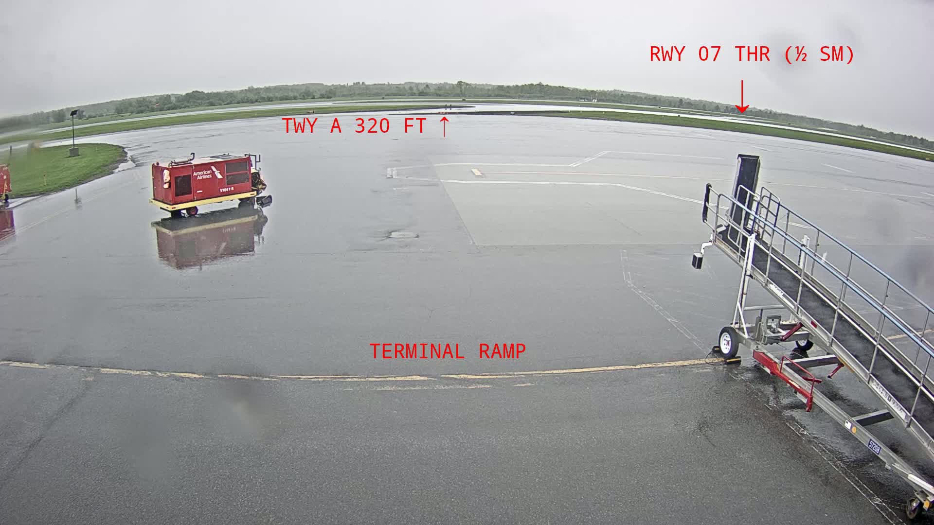 A wet airport tarmac shows a jet bridge, a baggage tug, and a runway in the distance under an overcast sky.