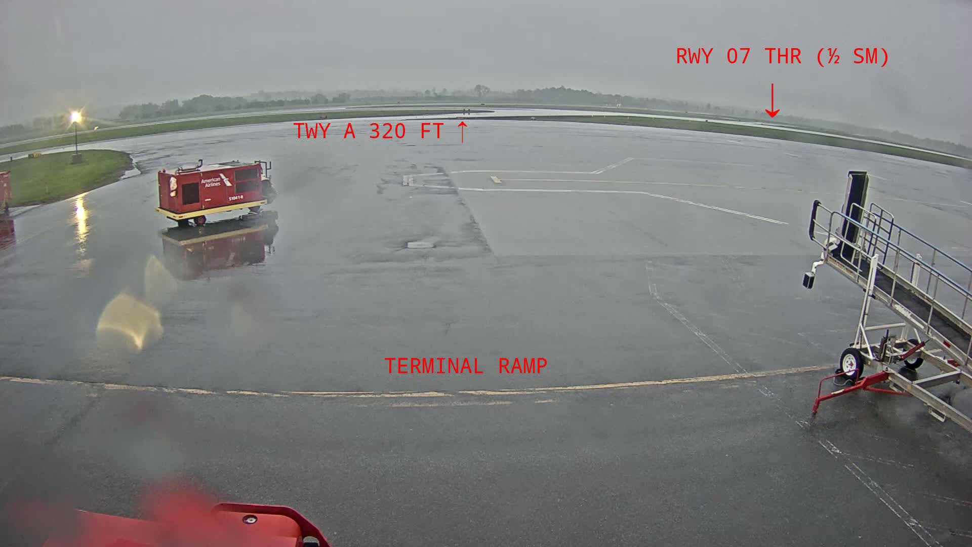 A wet airport tarmac is shown, with a red baggage vehicle and a jetway visible under an overcast sky.
