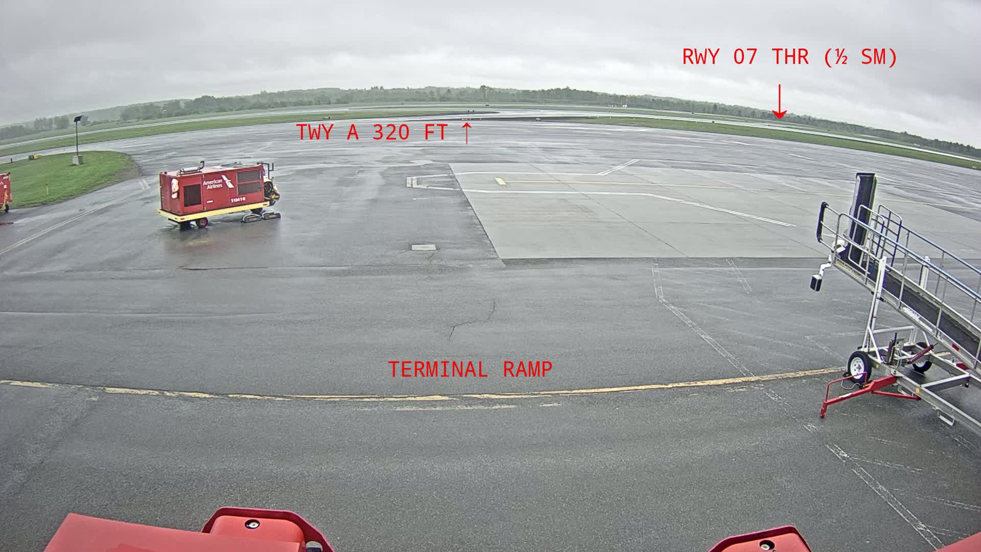 A wet airport tarmac is shown, with airport service vehicles and a jet bridge visible under an overcast sky.