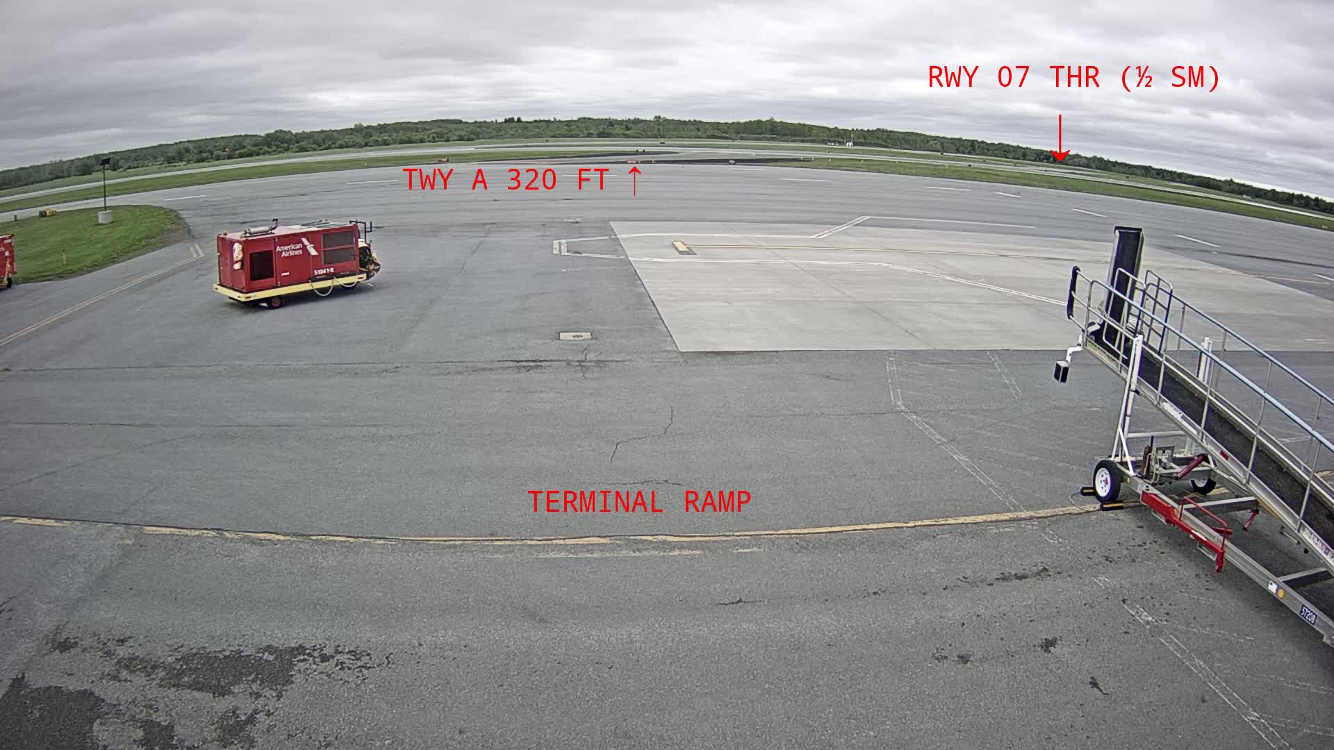 A tarmac view shows an airport terminal ramp, a baggage tug, a jet bridge, and a runway in overcast conditions.