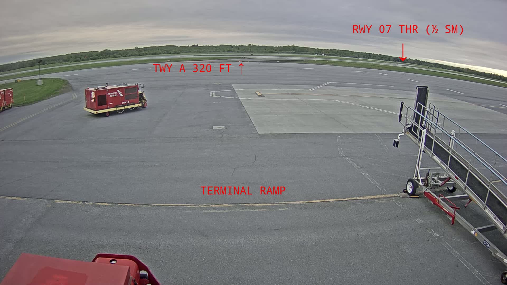 An airport tarmac under an overcast sky shows a baggage tug, a jet bridge, and a runway in the distance.
