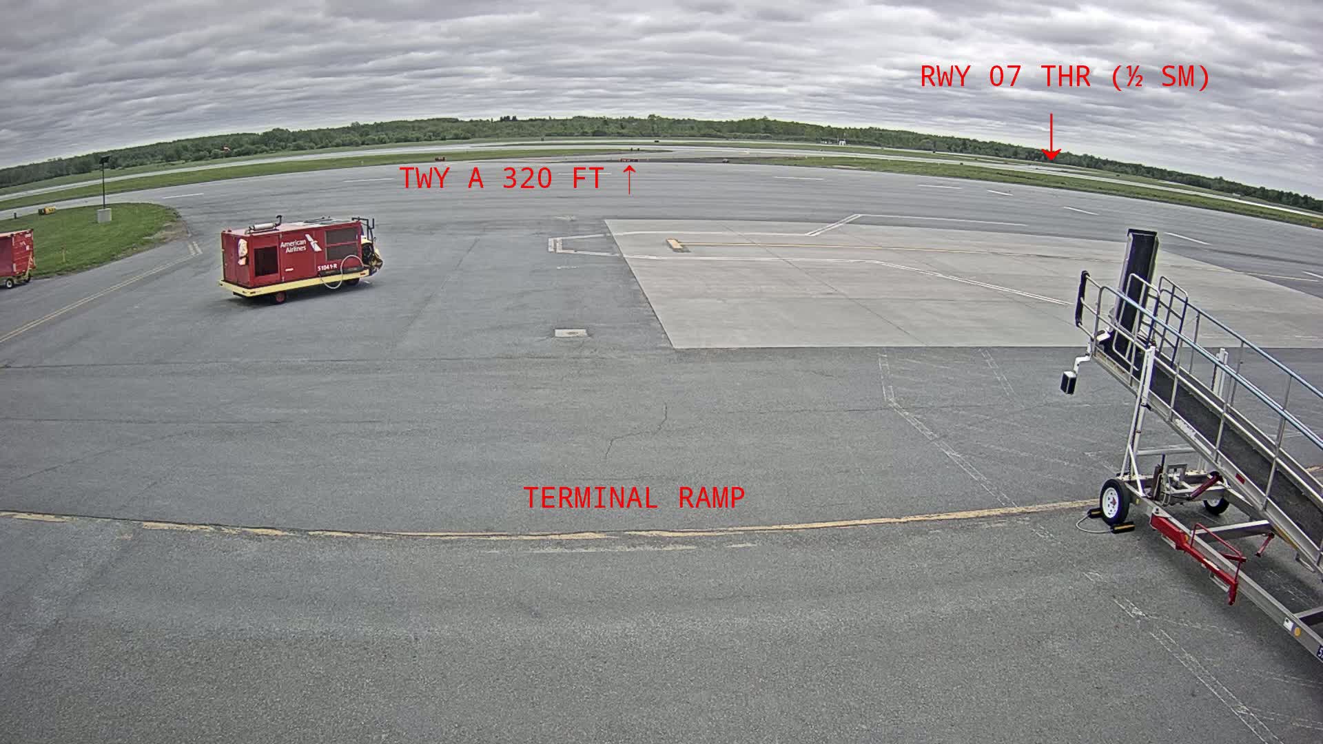 An airport tarmac under cloudy skies shows a baggage cart, a jet bridge, and a runway in the distance.