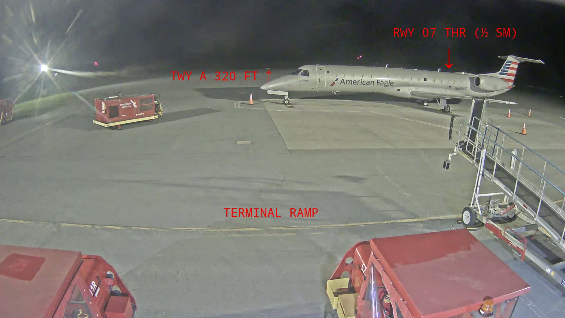 At night, an American Eagle plane is parked on a tarmac near a jet bridge and several pieces of ground support equipment.