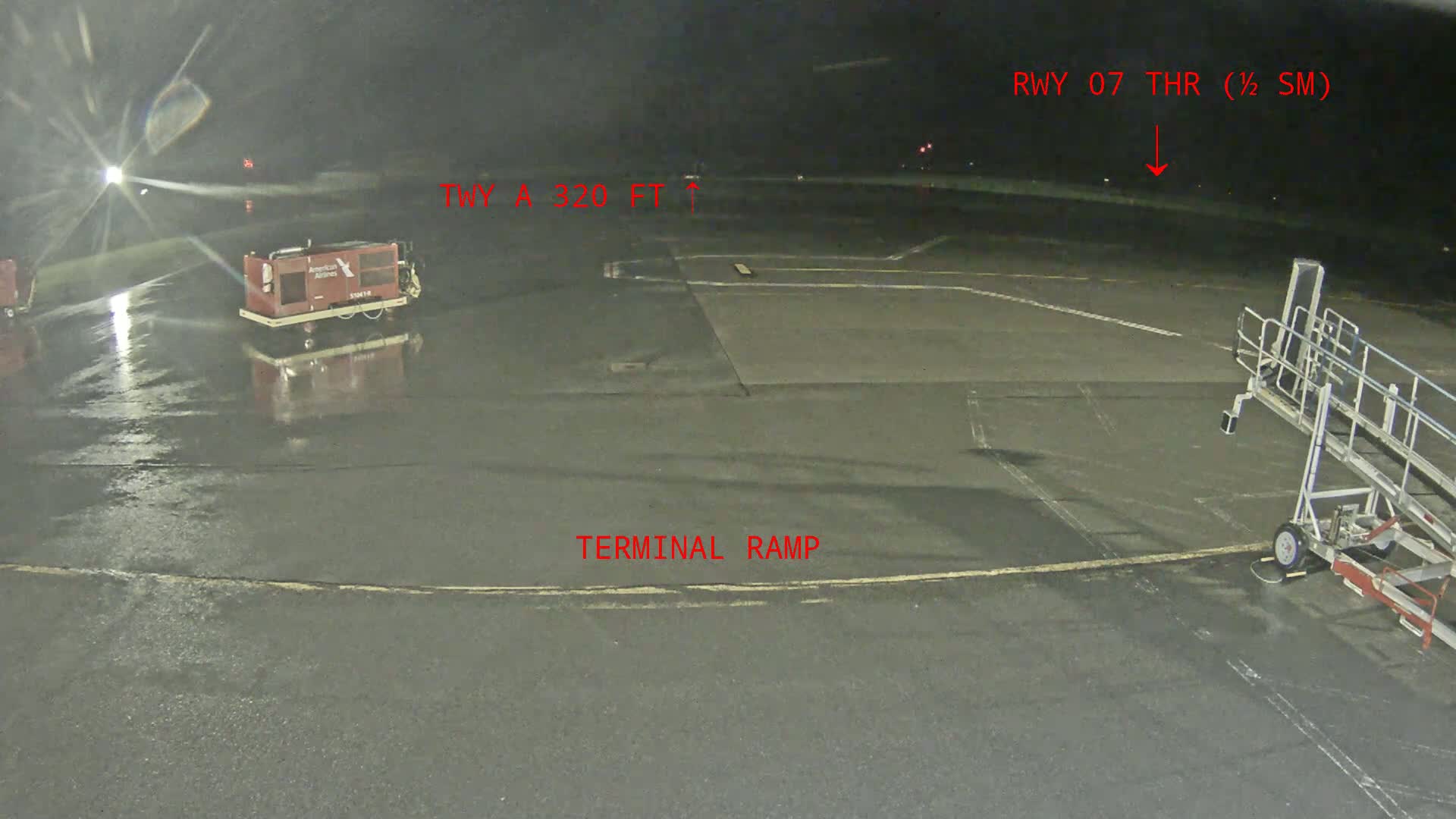 A wet airport tarmac at night shows a baggage cart and jet bridge near a terminal ramp.