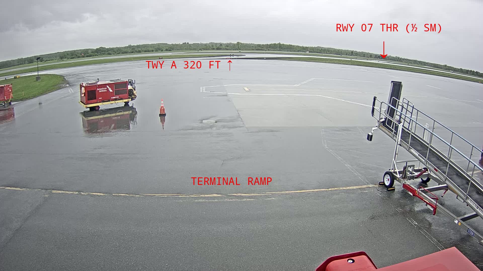 A wet airport tarmac is seen with a baggage tug, jet bridge, and a traffic cone; the background shows overcast skies and greenery.