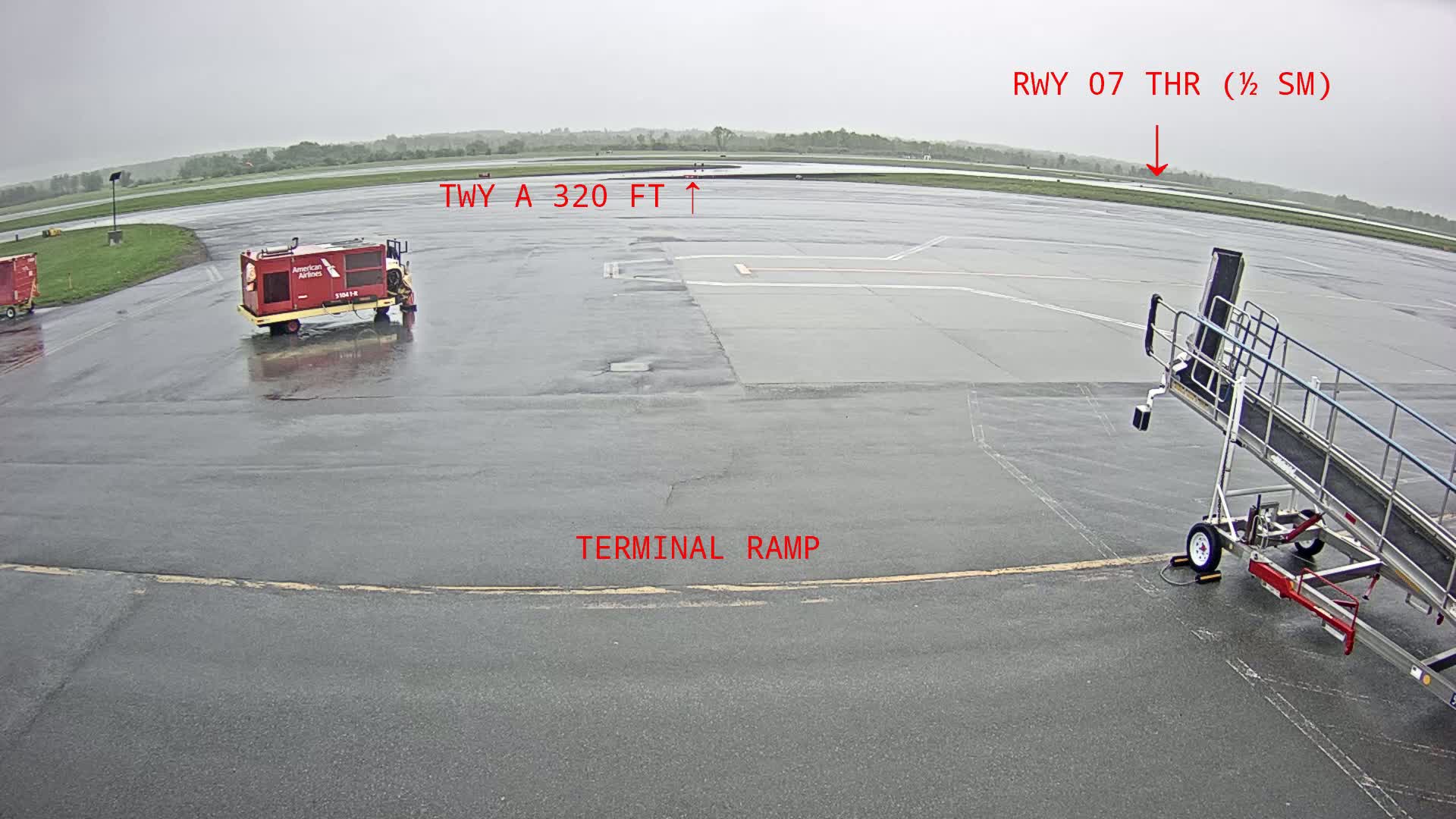 A wet airport tarmac shows a baggage tug, a jetway, and other airport equipment under an overcast sky.