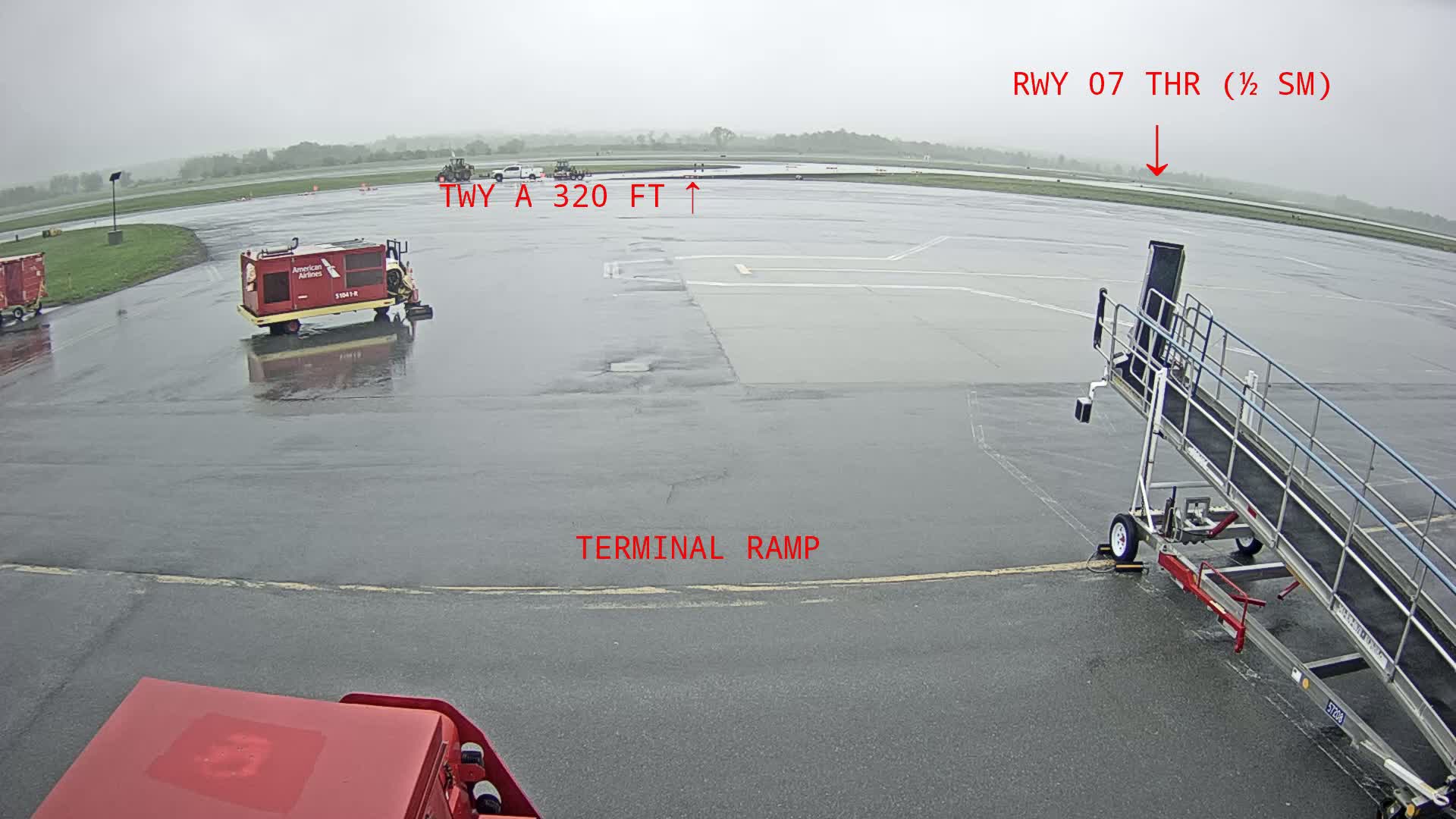 A wet airport tarmac is shown with service vehicles, a jetway, and a runway in the distance under an overcast sky.