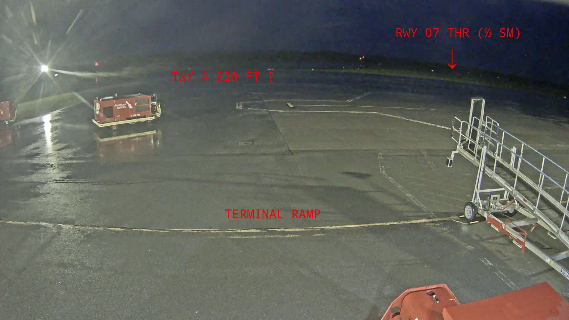 A wet airport tarmac at night shows a baggage cart, a jet bridge, and other airport equipment under dim lighting.