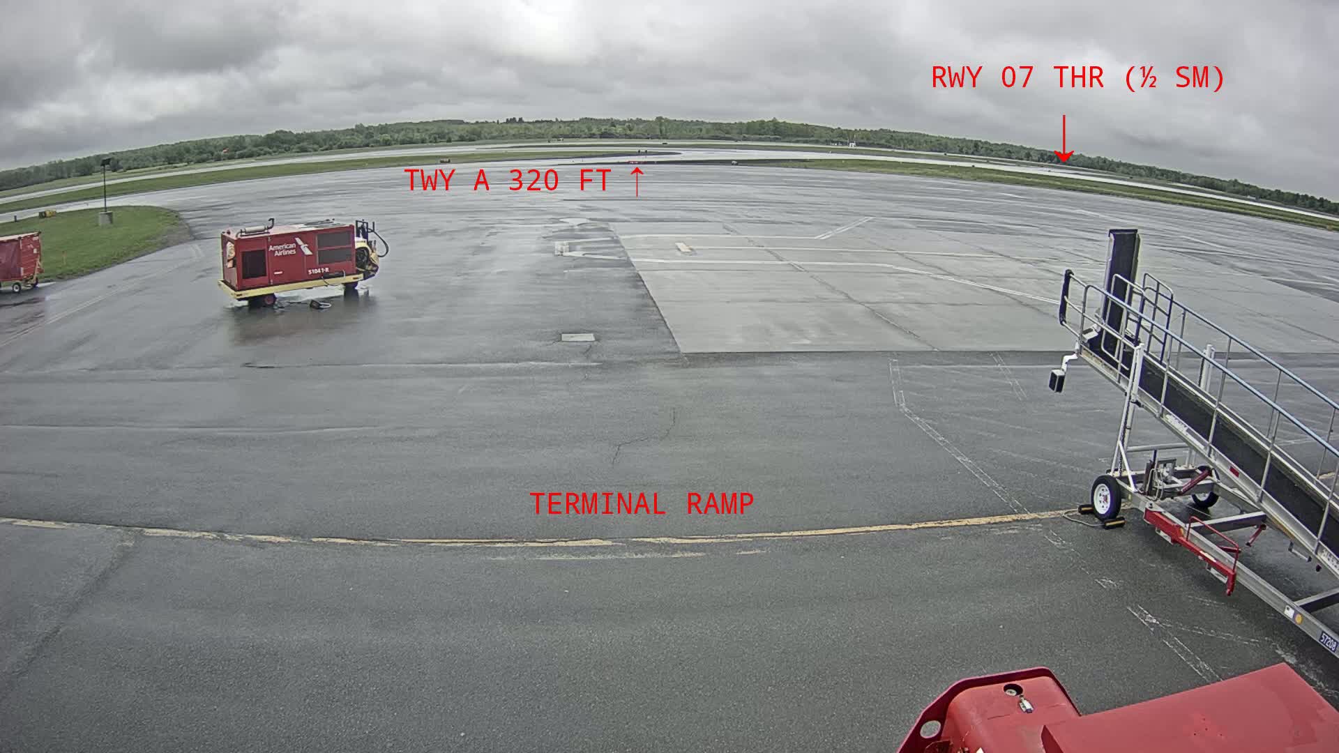 A wet airport tarmac is shown under an overcast sky, with various airport support vehicles and a jet bridge present.