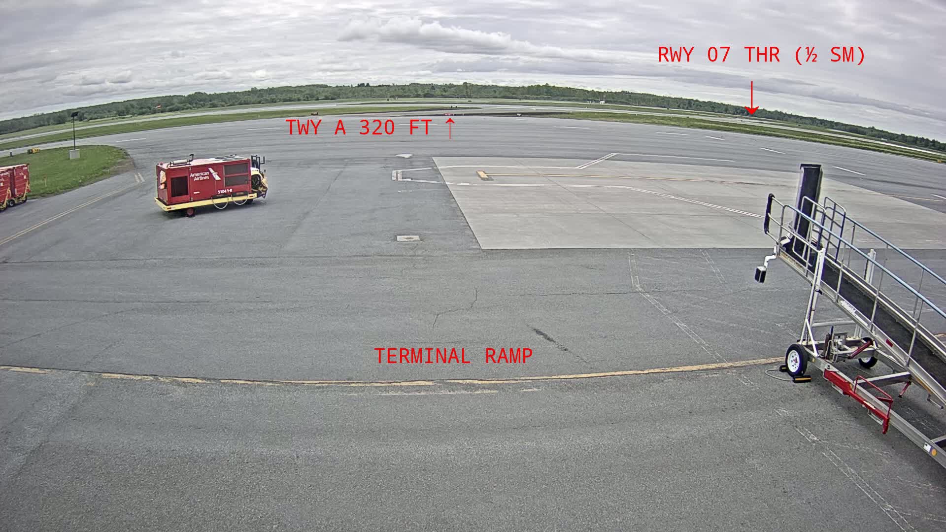 An airport tarmac on an overcast day shows a baggage tug, a jet bridge, and a portion of a runway in the distance.