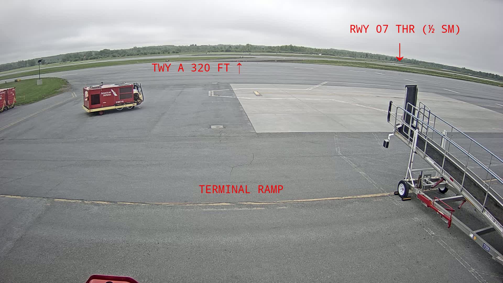 An airport tarmac under an overcast sky shows an airplane jet bridge, service vehicles, and a runway in the distance.