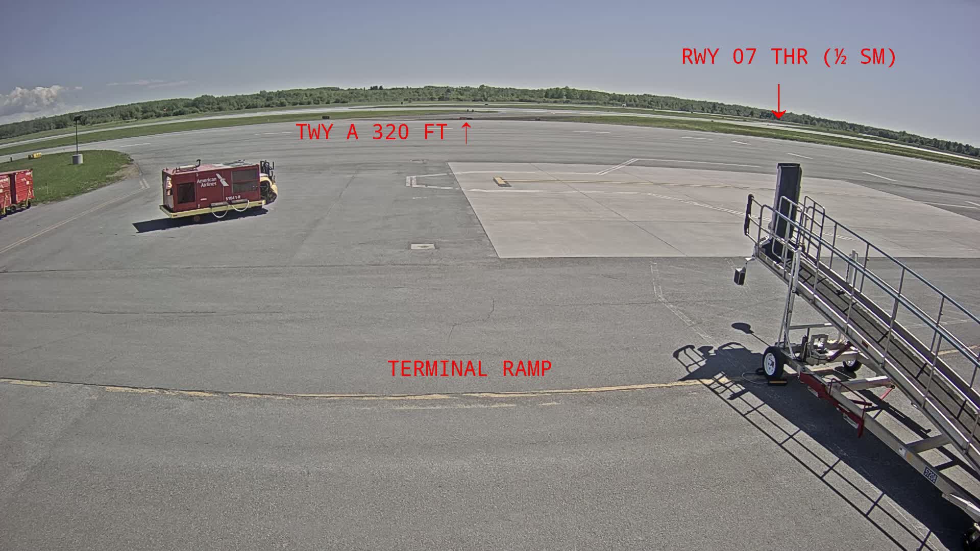 An airport tarmac on a sunny day shows a baggage cart, jet bridge, and other ground support equipment with a runway and grassy area visible in the distance.
