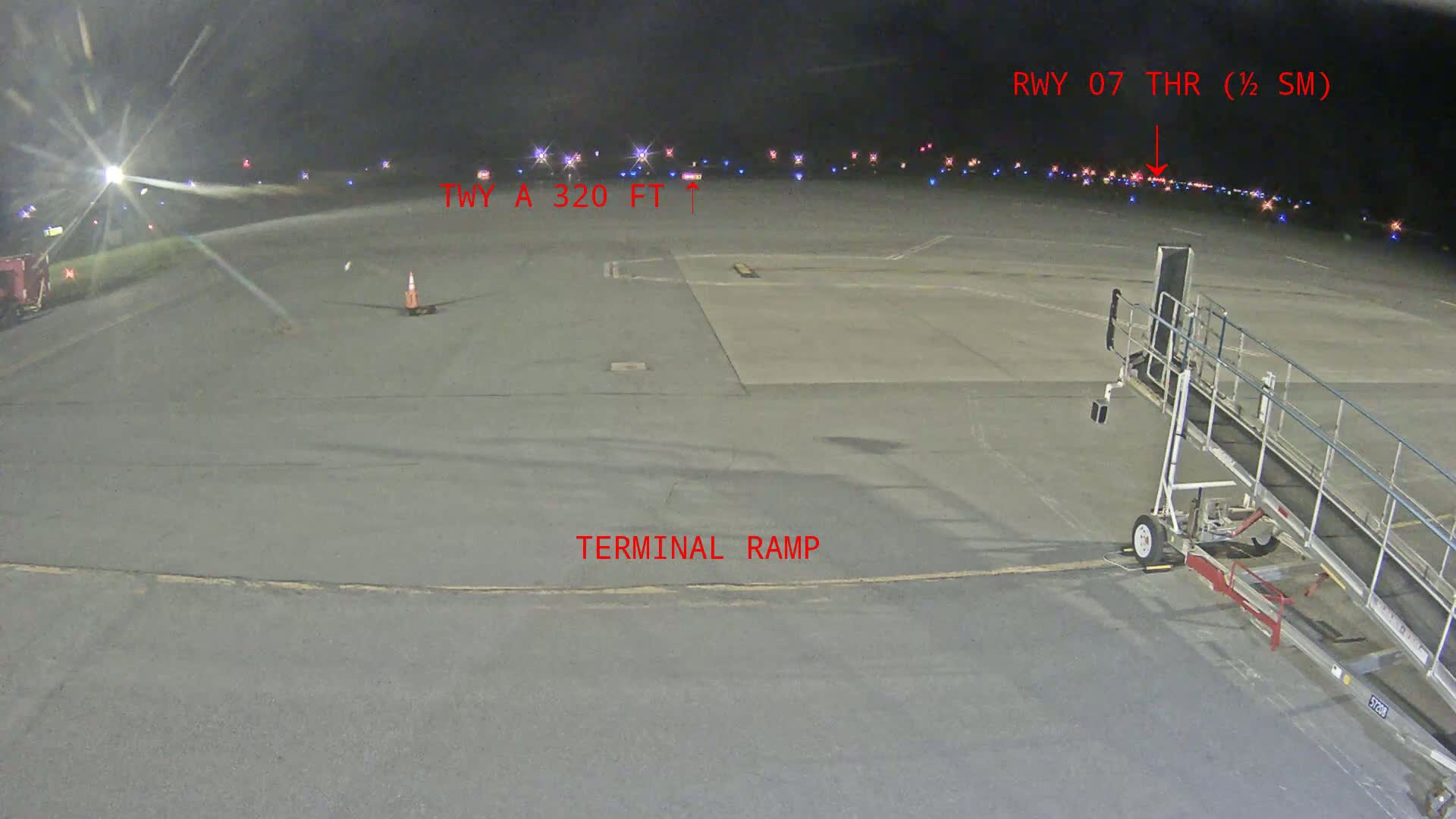 A nighttime view of an airport tarmac shows a jet bridge near a terminal ramp, with runway lights visible in the distance under a dark sky.