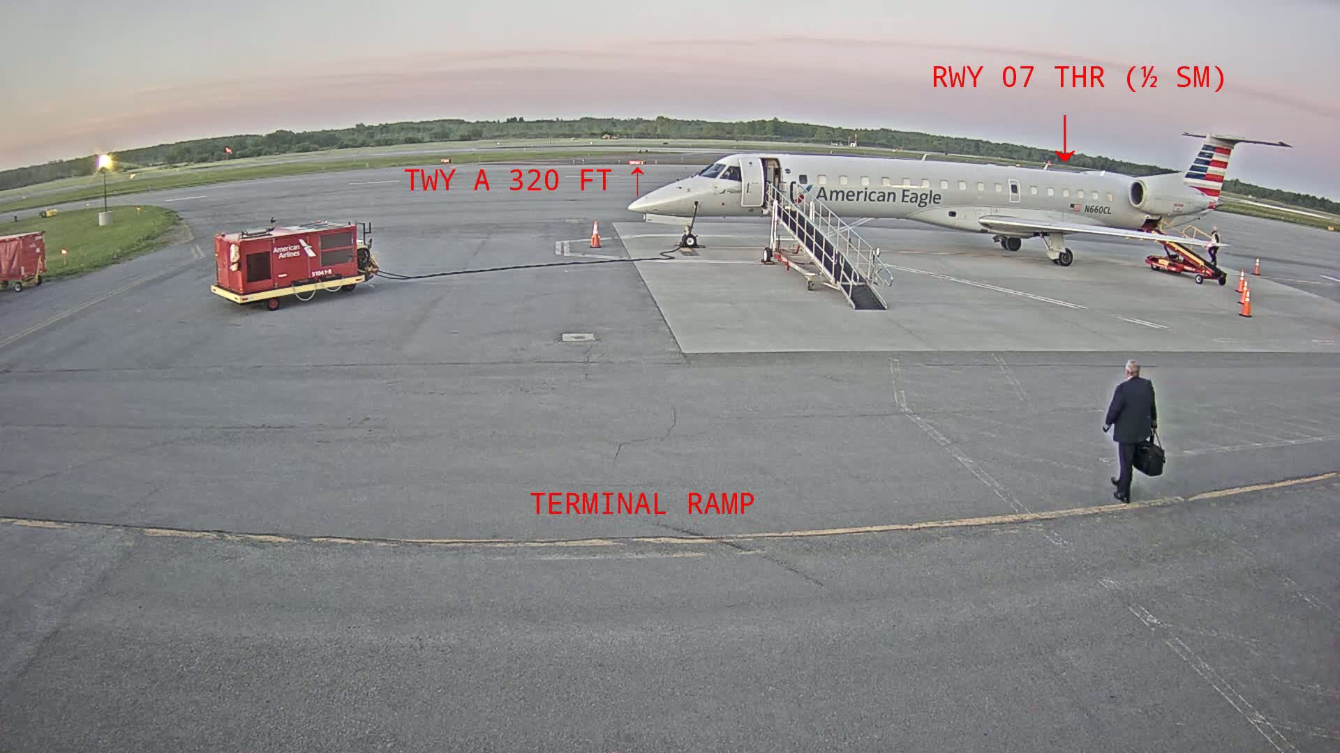 An American Eagle airplane is parked at a terminal ramp at an airport on a partly cloudy morning, with a man walking towards the terminal.