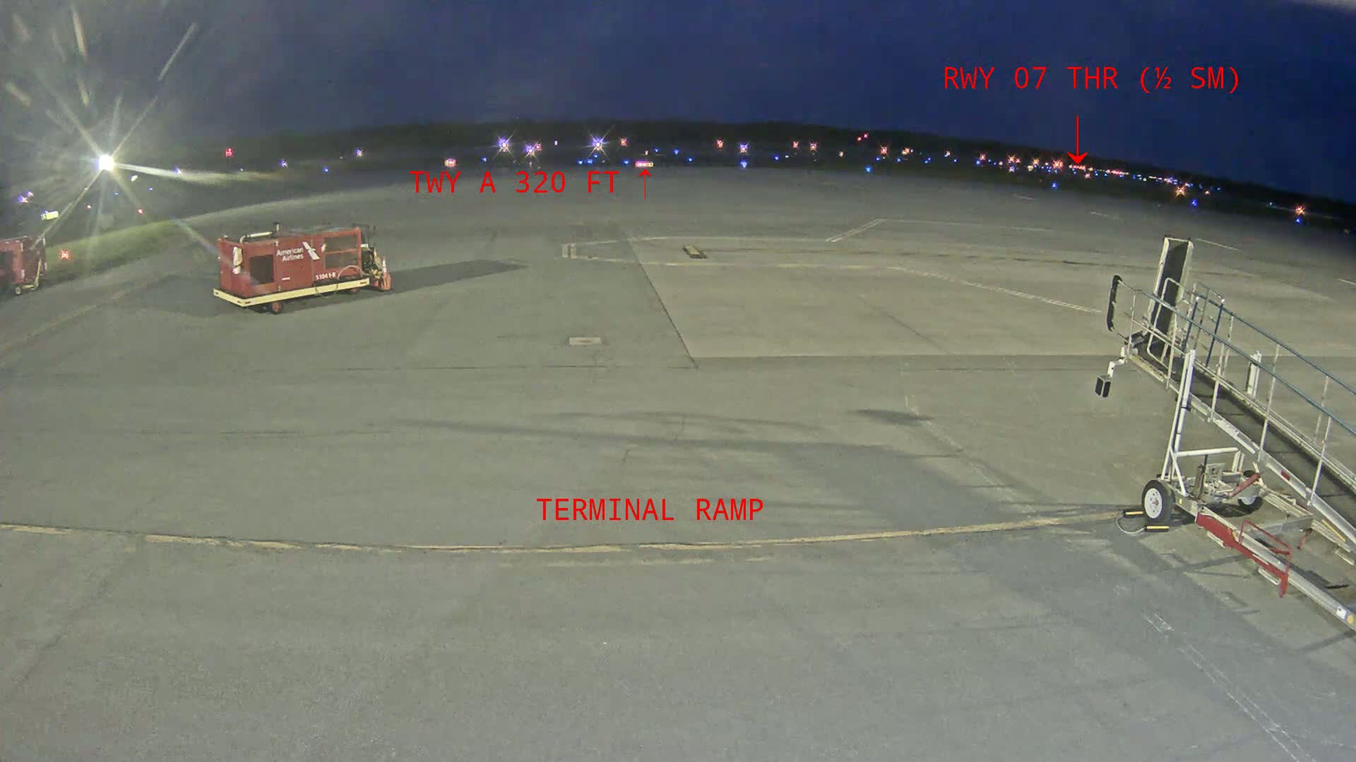 A dark tarmac airport scene at night shows an airplane boarding ramp, service vehicles, and runway lights in the distance.