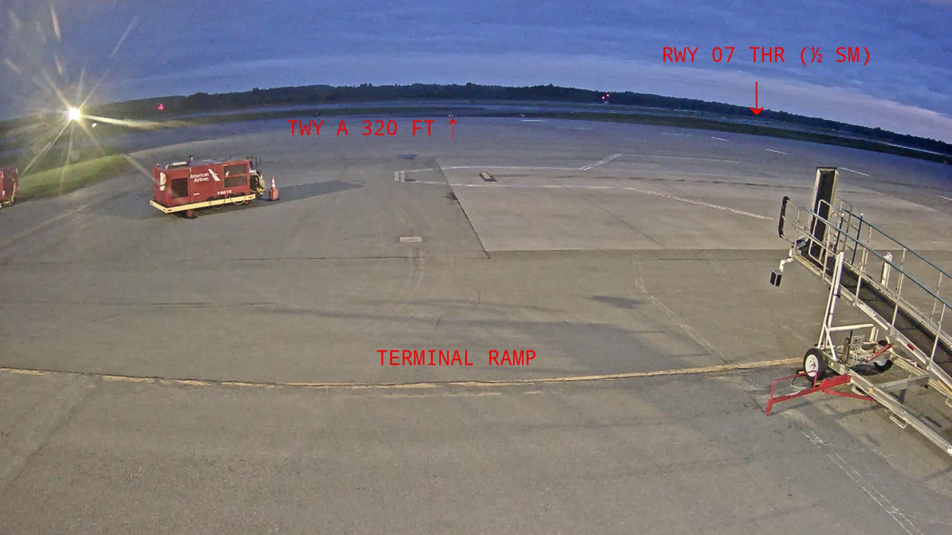A tarmac at dusk shows a red baggage cart, a jetway, and a runway in the distance.