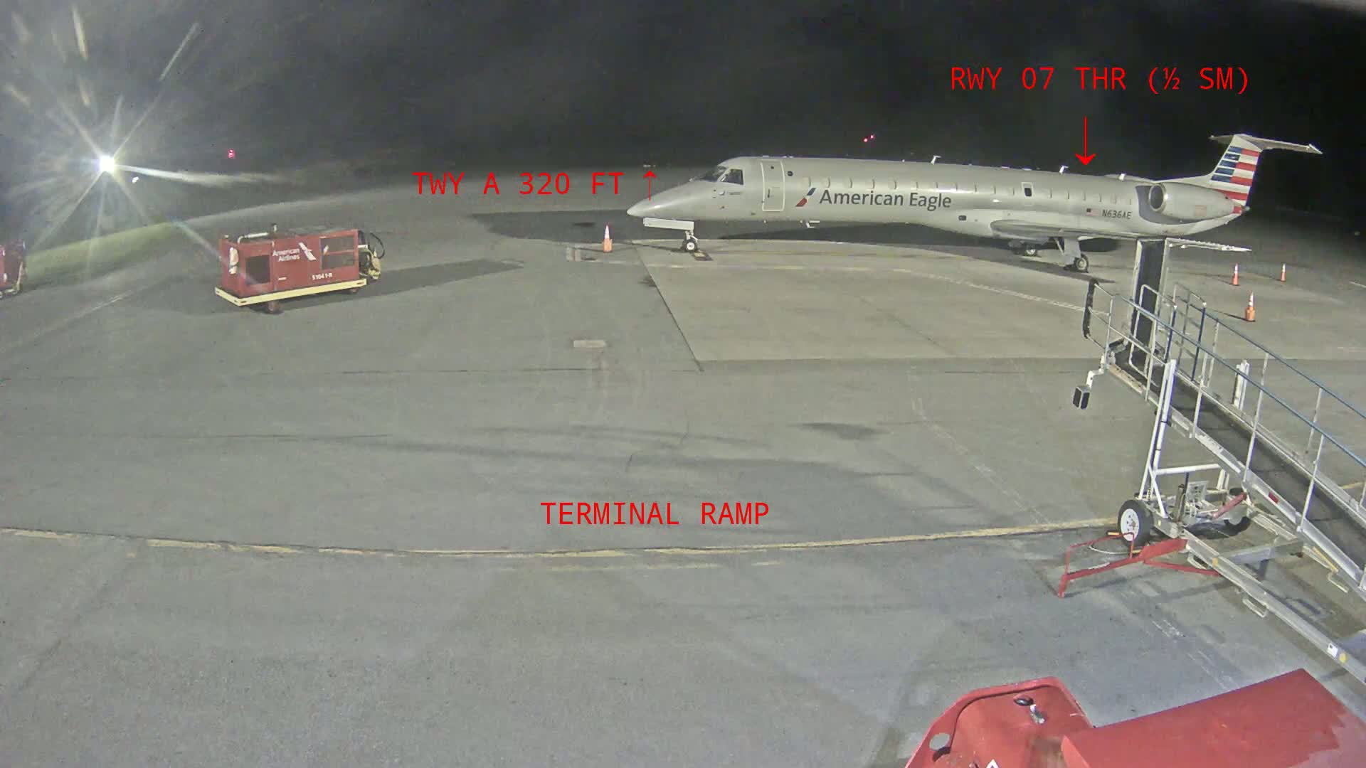 An American Eagle regional jet is parked on a tarmac at night near a terminal ramp and baggage cart.
