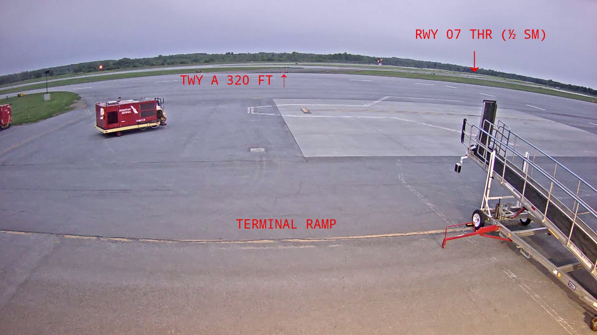 An airport tarmac under an overcast sky shows a baggage cart, a jet bridge, and a ground service vehicle.