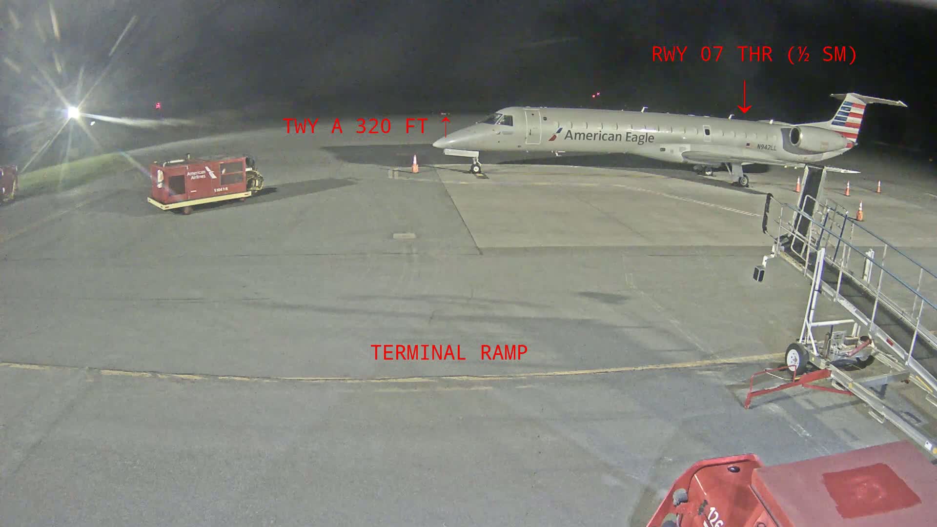 At night, an American Eagle airplane is parked on an airport tarmac next to a jet bridge, with ground support equipment nearby.