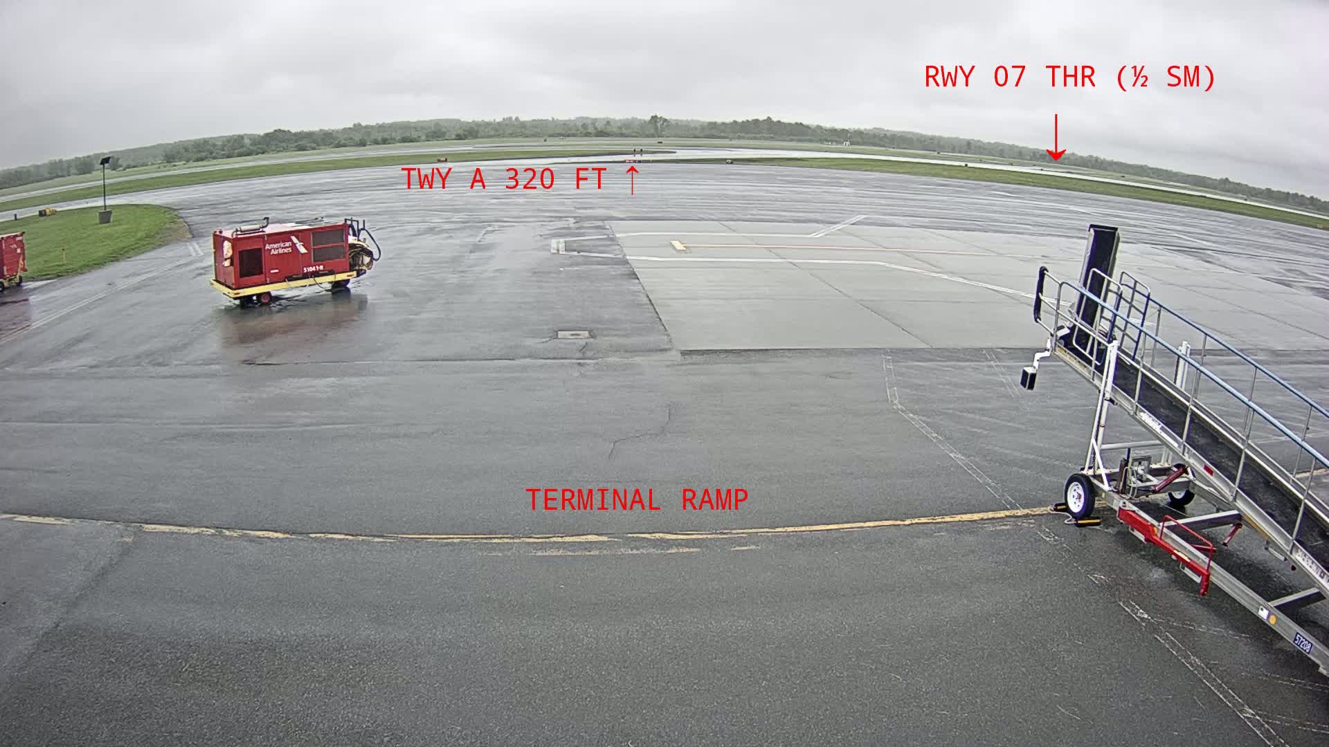 A wet airport tarmac is shown with a jet bridge, ground support equipment, and a view of a runway in the distance under an overcast sky.