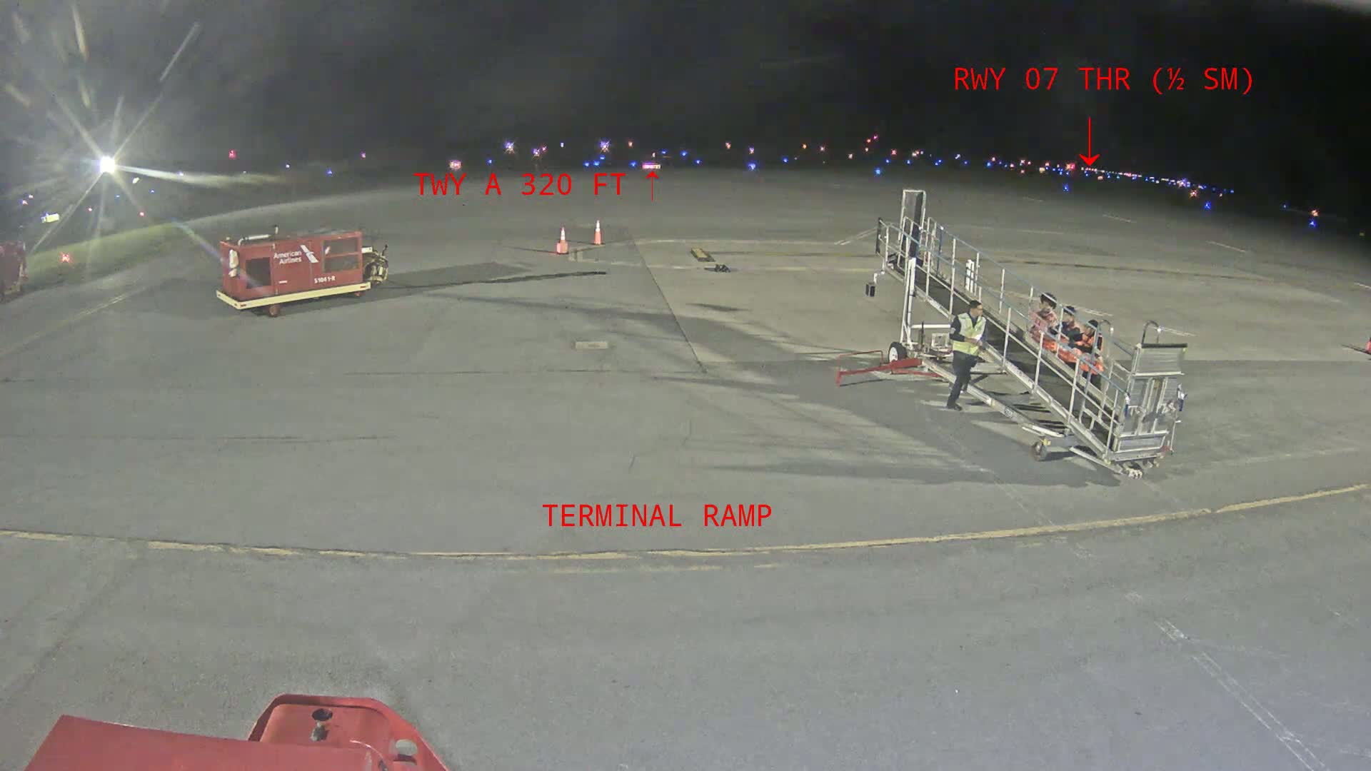 At night on an airport tarmac, a jet bridge is being used, with a baggage cart and other airport equipment nearby, under dark, clear skies.