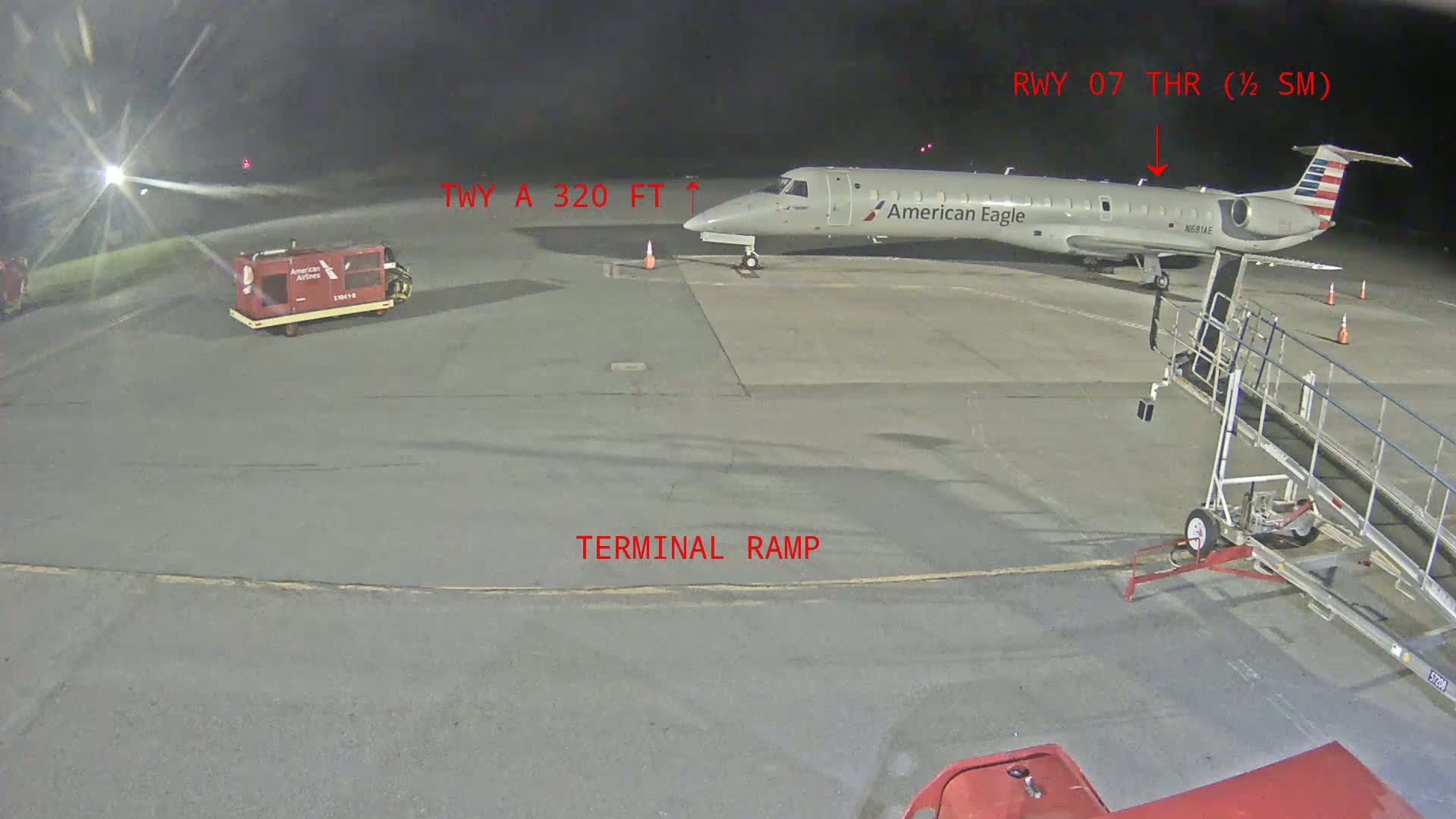 At night, an American Eagle regional jet is parked at an airport terminal gate, with a baggage tug nearby and a jet bridge attached.