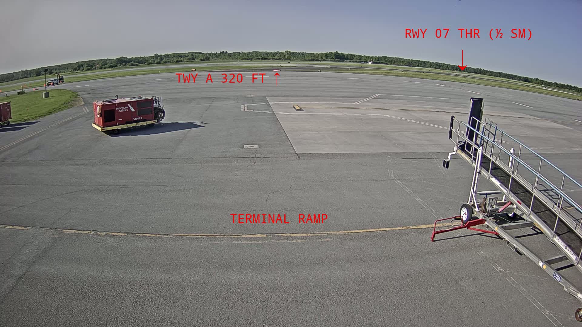A terminal ramp at an airport on a sunny day shows a baggage cart and a jet bridge, with a runway visible in the background.