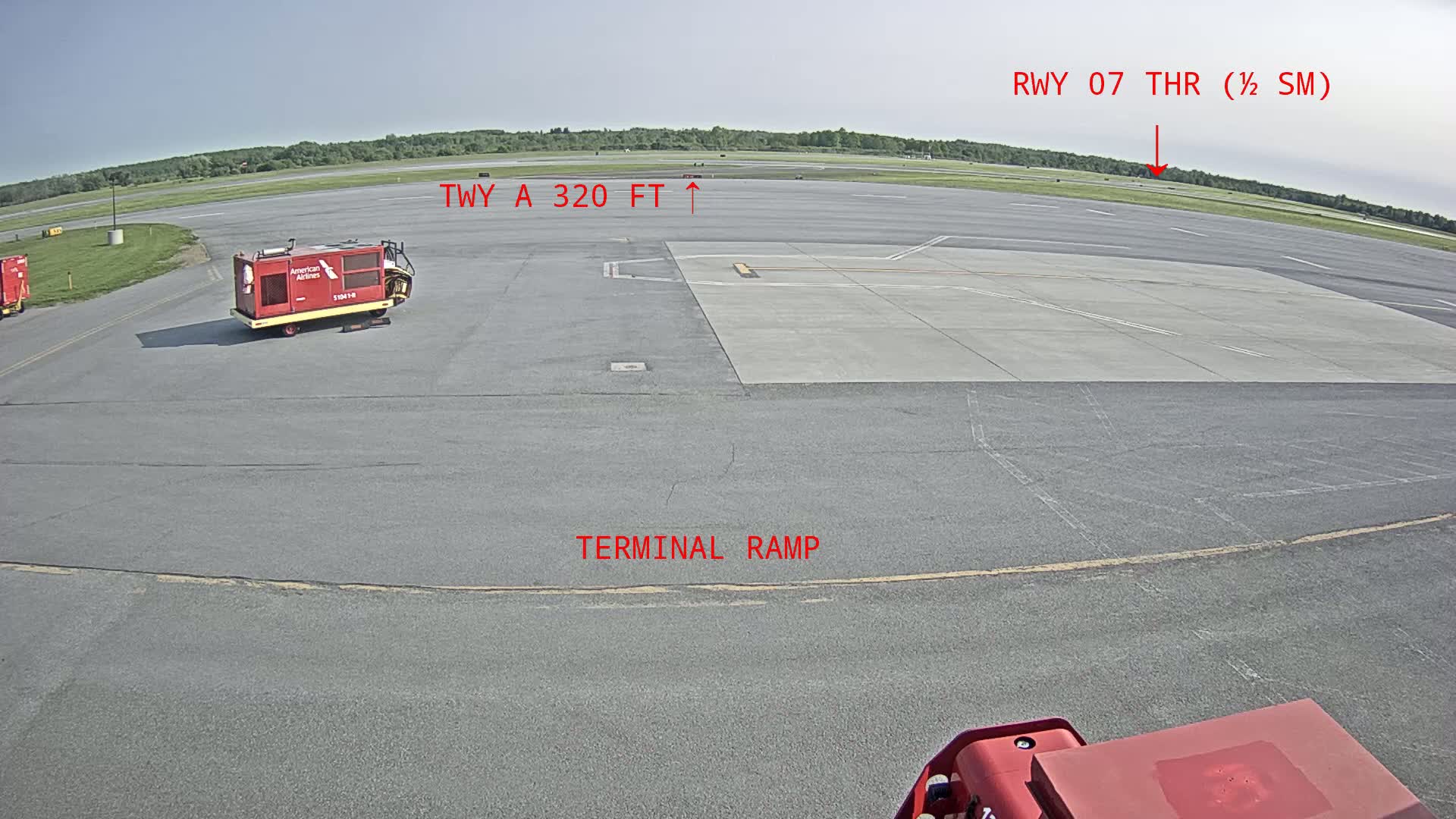 A tarmac at an airport on a sunny day shows a terminal ramp with two pieces of ground support equipment and a runway in the distance.
