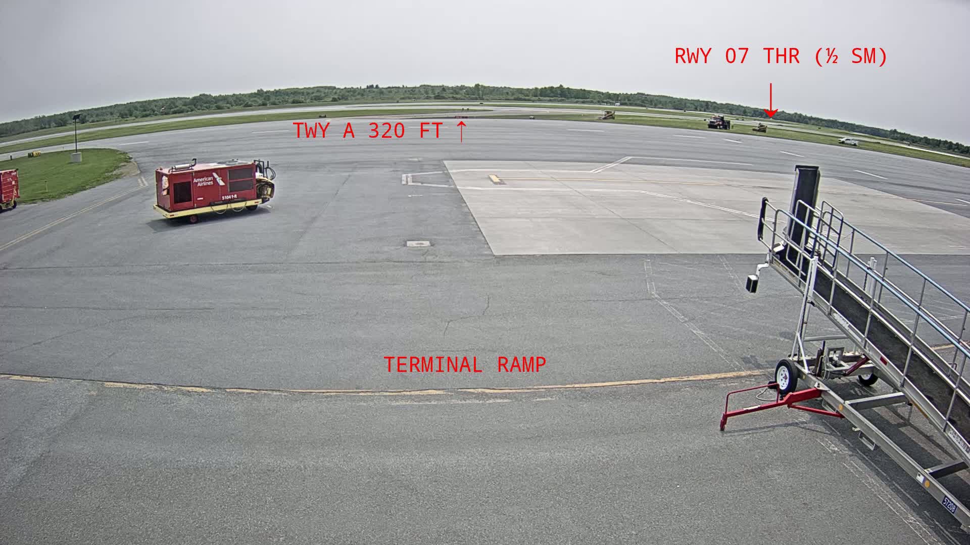 A mostly empty airport tarmac on a partly cloudy day shows a baggage tug, a jet bridge, and a few other vehicles.
