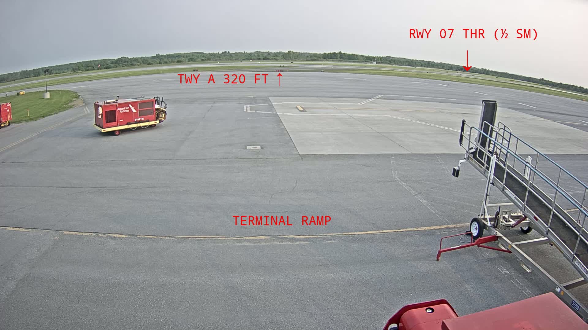 An airport tarmac under an overcast sky shows a jet bridge, several ground service vehicles, and a runway in the distance.