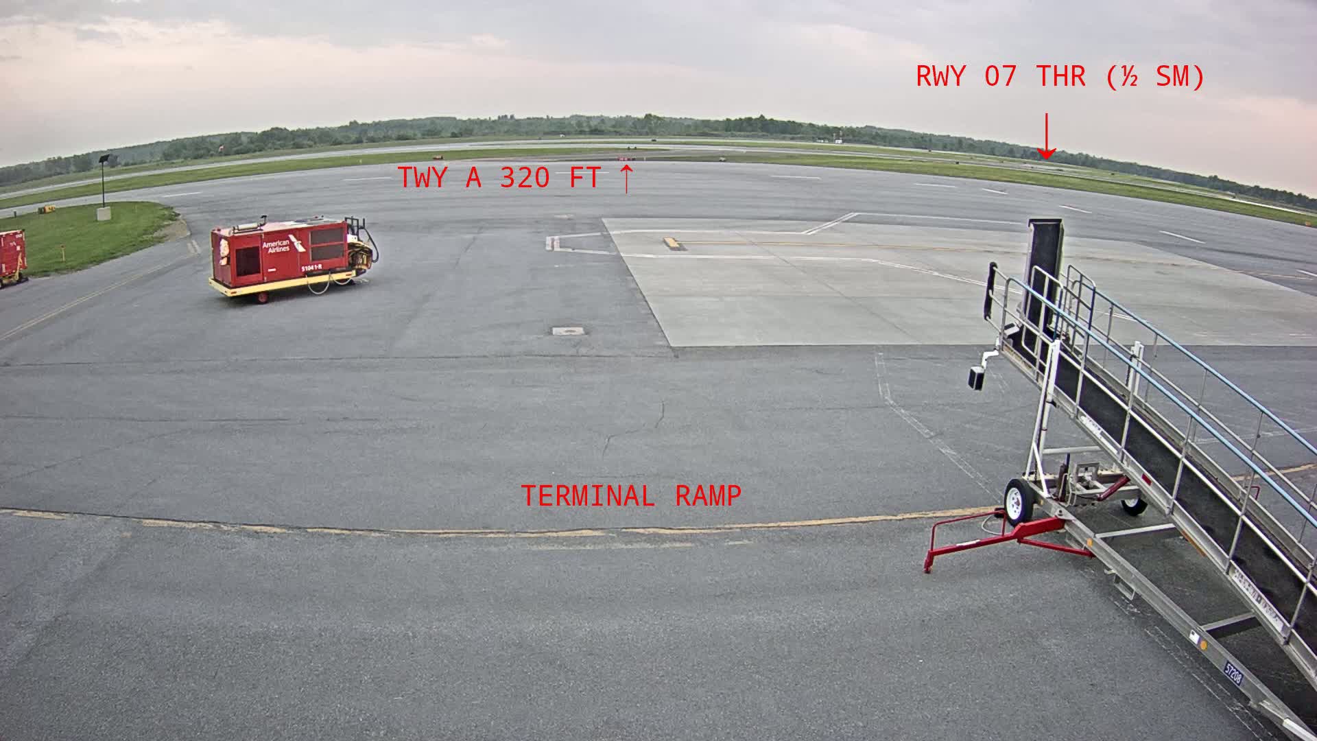 An airport tarmac under an overcast sky shows a baggage cart, a jet bridge, and ground support equipment.