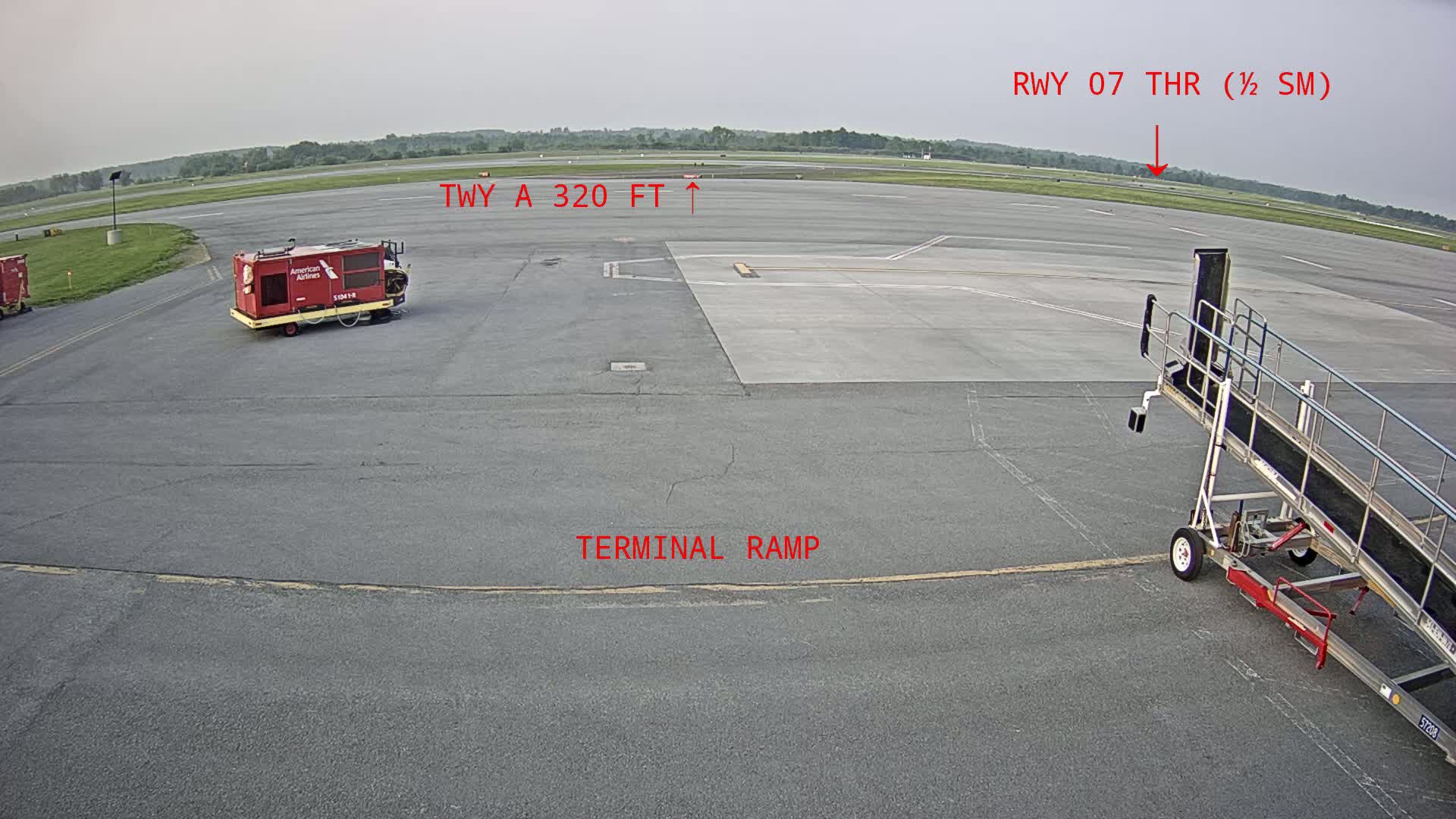 A mostly clear, overcast day at an airport tarmac shows a baggage cart, a jet bridge, and some airport ground support equipment near a terminal ramp.