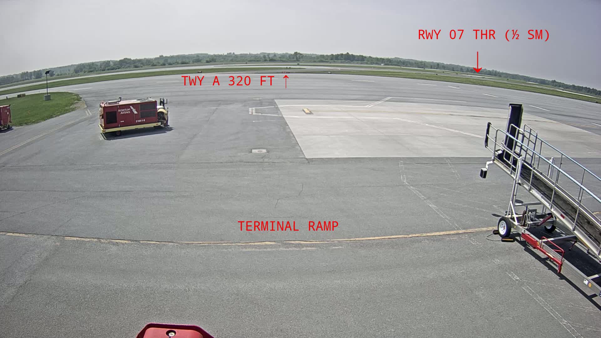 An airport tarmac on a sunny day shows a baggage cart, a jetway, and a runway in the distance.